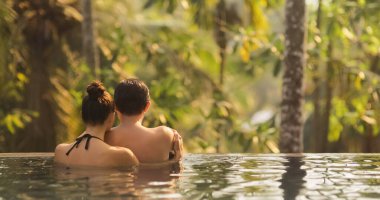 Panoramic photo of young adult couple in love swimming together in open-air infinity pool enjoying tropical vacation on villa looking away at rainforest on sunset. High quality photo