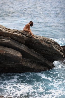 Güzel bir bikinili kadın modelinin hava drone görüntüsü. Kayalık bir uçurumda oturup denize bakan inanılmaz ince bir vücudu var. Fırtınalı dalgalar ve muhteşem bir kadın modelin olduğu destansı bir sahne. Yüksek kalite