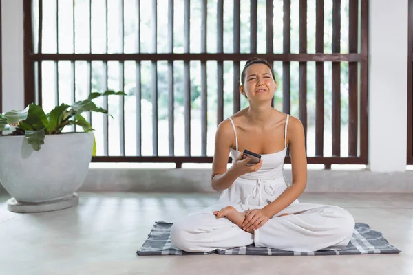 Sad yoga beginner woman using a mobile phone during yoga educational practice indoors. Fit young adult female crying because of a painful pose. High-quality photo
