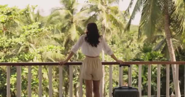 トロリーバッグ付きの別荘のバルコニーに立つ若い観光客の女性のスローモーション映像 到着後の熱帯雨林の美しい景色を撮影 — ストック動画