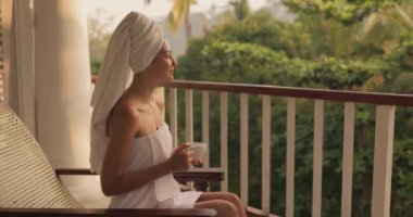 Açık bir tatil beldesinin balkonunda dinlenen beyaz havluyla bir fincan kahve veya çay içen mutlu bir kadın tropikal bir ülkede, bir yağmur ormanına karşı bir koltukta oturuyor. Yüksek