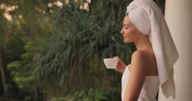 Mükemmel bir sabah. Açık bir tatil beldesi terasında tropik bir ülkede palmiye ağaçlarına karşı durduktan sonra beyaz havluyla bir fincan kahve ya da çay içen mutlu bir kadının yakın plan portresi. Kenar