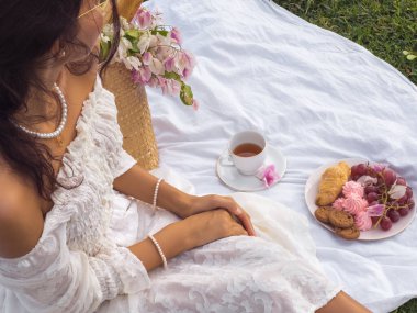 Genç bir kadın şık bir şekilde parkta romantik bir pikniğin tadını çıkarıyor. Klasik detaylar, boho battaniyesi, çay fincanı, bir tabak tatlı ikram..