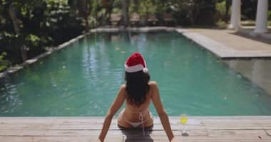 Mayolu ve Noel Baba şapkalı bir kadın havuzun arka tarafında oturuyor, kış tatilinin tadını çıkarıyor ve yeni yılı tropik bir yerde kutluyor. Bir kadın ortalarında elini kaldırdı.