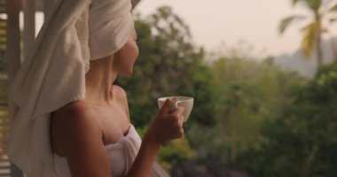İyi sabahlar. Açık bir tatil beldesinin balkonunda, ormanın önünde, mükemmel bir sabahta, beyaz havluyla bir fincan kahve ya da çay içen mutlu bir kadının yakın plan portresi.