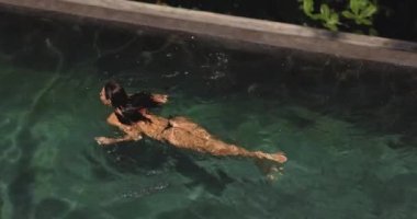 Siyah bikinili zayıf, genç ve seksi bir kadının lüks bir tropikal havuzda yüzüşünün insansız hava aracı görüntüsü. Yavaş çekim 4k görüntü