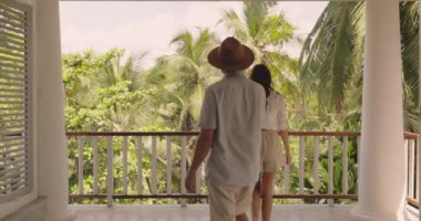 Tramvay Çantalı Genç Turist Çifti Tropikal Villa Oteli 'ne kayıt yaptırıyor, Yaz Tatili boyunca Balkondan Palmiye Ağaçlarıyla Bereketli Yeşil Orman' da Romantik Manzaranın tadını çıkarıyor