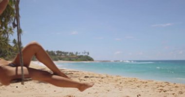 Tropik tatil konsepti. Siyah mayo giymiş şık, ince bikinili bir plaj kadını yaz tatillerinde tropikal deniz kıyısında palmiye ağacında salıncakta sallanırken eğleniyor. Yüksek kalite kavramsal 4k yavaş
