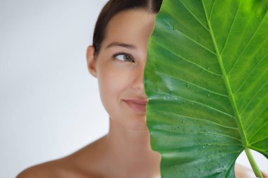 Parlak, kusursuz genç kadın çevre dostu güzellik salonu ürünlerini tanıtıyor. Yeşil bir bitkisel yaprağın arkasında yüzünü örten güzel bir kadının yakın plan portresi.