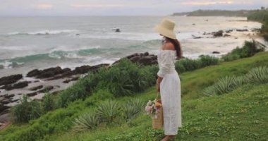 Romantik kadın denizi kıyısındaki boho pikniği için şık bir yaz plajı kıyafeti. Şık, şık, modaya uygun beyaz bir elbise içinde, klasik aksesuarlar ve plaj çantasındaki çiçek buketi ile duruyor.