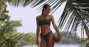 Tanned positive bikini beach woman, holding a fresh coconut in front of a tropical lagoon under a palm tree leaf, during summer sea vacation
