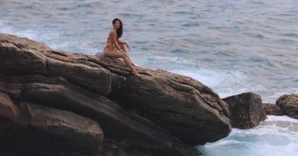 岩の崖に座って海を見ている素晴らしいスリムな体を持つ美しいビキニの女性モデル 嵐の波と豪華な女性モデルの壮大なシーン 高品質のスローモーション映像 — ストック動画