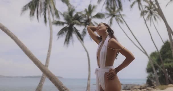 夏のトロピカルビーチでパームツリーでリラックスした白い水着の豪華なファッショナブルな日焼けした女性 — ストック動画