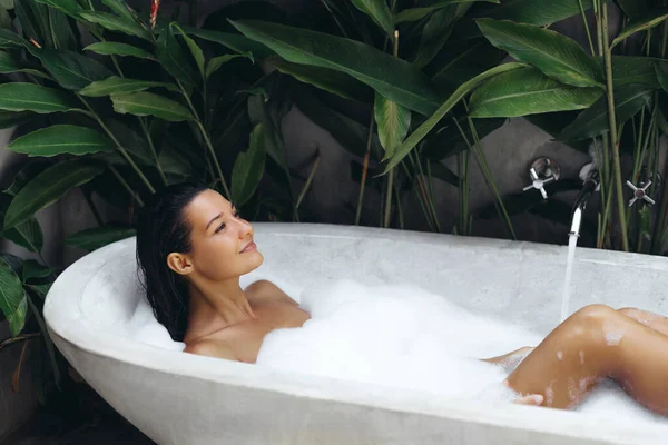 年轻女人躺在充满泡沫的浴缸里 一边吹泡泡一边玩乐 热带家庭温泉的概念 迷人的女士在浴池中放松 享受美丽和护肤的一天 — 图库照片