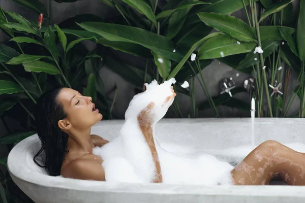 年轻女人躺在充满泡沫的浴缸里 一边吹泡泡一边玩乐 热带家庭温泉的概念 迷人的女士在浴池中放松 享受美丽和护肤的一天 — 图库照片