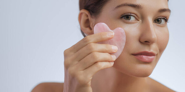 Beauty and skincare concept with a beautiful young adult woman doing a skincare routine, self-massage with the gua sha tool. Female model using a pink gemstone massager for facial lifting. Studio shot