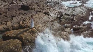 Uçurumun kenarında durup denize bakan beyaz elbiseli güzel bir gelinin hava aracı görüntüsü. Fırtınalı dalgalar ve muhteşem bir kadın modelin olduğu destansı bir sahne. Profesyonel 4k görüntüsü