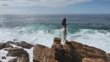 Uçurumun kenarında durup denize bakan beyaz elbiseli güzel bir gelinin hava aracı görüntüsü. Fırtınalı dalgalar ve muhteşem bir kadın modelin olduğu destansı bir sahne. Profesyonel ağır çekim.