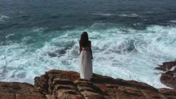 从空中俯瞰着一个美丽的新娘 穿着漂亮的白色连衣裙 站在悬崖上凝视着大海 一个史诗般的场景 波涛汹涌 还有一个漂亮的女模特 专业4K镜头 — 图库视频影像