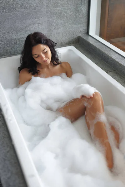 Bathing and daily routine concept. Front top view of attractive young adult woman model taking bath in luxury full of foam bathtub, washing her body, touching her silky skin on her leg. High quality