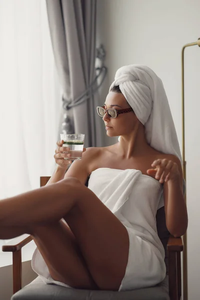 Young beautiful confident woman after bath or shower in sunglasses dressed in towel on her head. Sexy carefree model sitting on armchair, drinking in posh apartment or hotel room. High quality photo