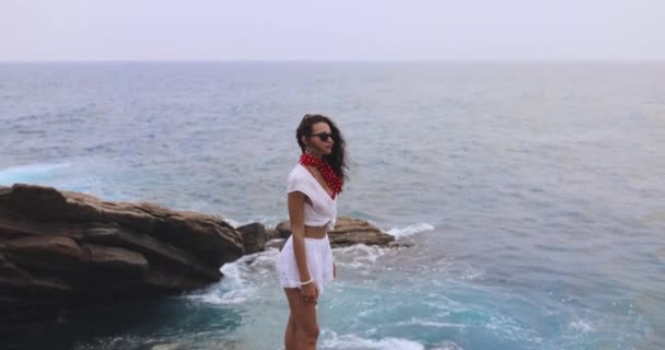 岩の崖の上に立って空を見ているサングラスの白い夏の服のスタイリッシュな女性 嵐の波 豪華なガールモデルと壮大なシーン — ストック動画