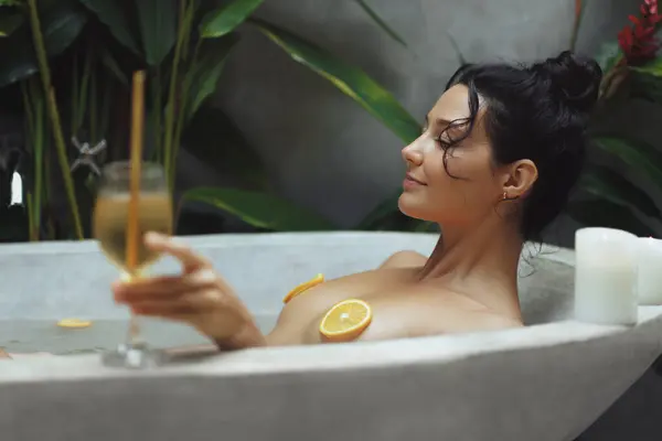 Hotel spa relax and skin whitening beauty portrait of smiling young female model resting in bathtub full of oranges and lemons with closed eyes drinking champagne. Attractive naked millennial woman