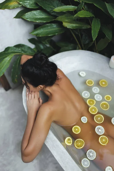 Spa relax of naked young adult female model resting in concrete bathtub full of oranges and lemons slices. Rear view of attractive bare millennial tanned woman relaxing, bathing in clear water, modern