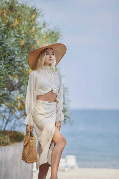 Stunning rich blonde woman in luxury summer beach outfit relaxing outdoors against sea resort. A fashionable romantic young adult lady wearing a trendy straw hat, white blouse, and skirt, standing at