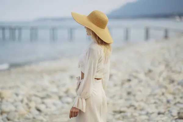 Stunning blonde woman in summer beach outfit relaxing outdoors against sea background. Back view of fashionable romantic young adult lady wearing a trendy vintage straw hat, white blouse, and skirt