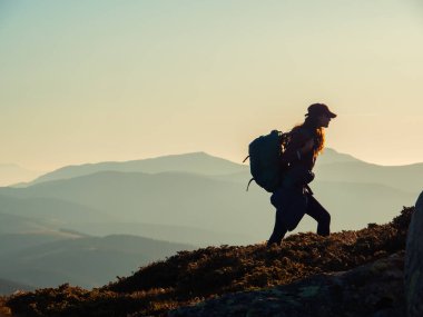 Dağ yürüyüşü sırasında sırt çantalı gezgin bir kadının silueti. Arka planda akşam dağlarının görkemli manzarası.
