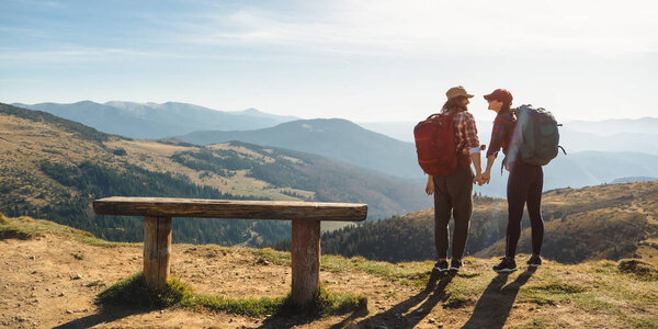 Couple of hikers with backpacks enjoying valley landscape view from top of a mountain. Happy Young adult tourists, man and woman hugging and laughing. Panoramic view of mountain hills, Carpathian
