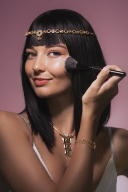 Kadın makyaj fırçası kullanarak yüzüne pudra sürüyor. Kleopatra tarzı moda modeli yanaklarına allık sürüyor. Güzellik kozmetikleriyle yüzleş ve akşam kavramsal fotoğraf yap