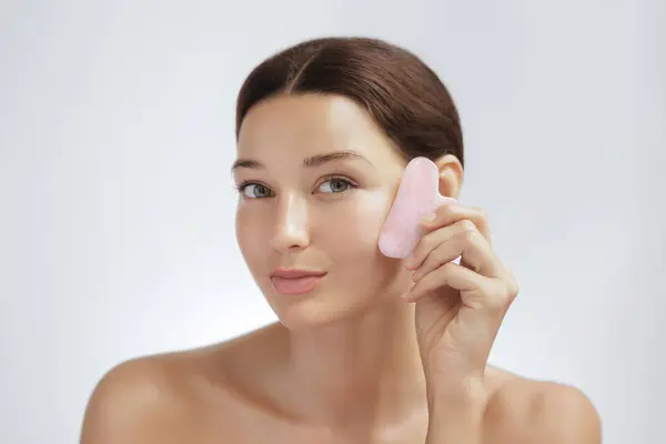 Beauty face care. Young Adult woman doing face self-massage with pink jade for spa skin care treatment at home. Girl model using natural massager tool portrait on blue background