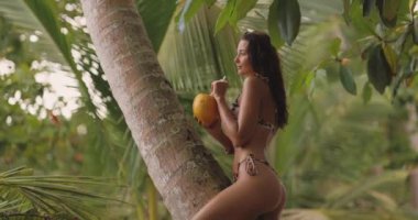 Genç, çekici bir Asyalı kadın model tropikal deniz kıyısında seksi leopar desenli bikini ve hindistan cevizi içerken kaygısız bir gülümseme giyerek mükemmel bir günün tadını çıkarıyor.