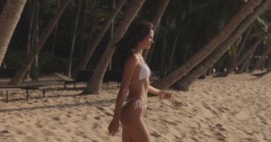 Beyaz mayo giymiş şık, bronzlaşmış bir kadının yaz tropik plajda palmiye ağaçlarıyla dinlendiği lüks tatiller..