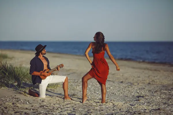 官能的なロマンスを愛する幸せなカップル 日没にビーチで踊る赤いドレスでウクレレのセレナーディングと女の子を果たしている白人男性 髭を生やしたハンサムな男と見事な女持っていますロマンチックな入札 — ストック写真