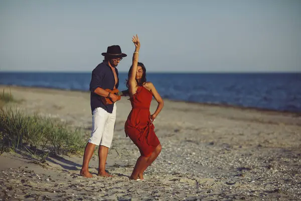 恋爱中的快乐夫妻 日落时分 一个白人男子在沙滩上弹奏着小夜曲 一个身穿红衣的女孩在沙滩上跳舞 一个留着胡子的英俊男人和迷人的女人有着浪漫的温柔 — 图库照片