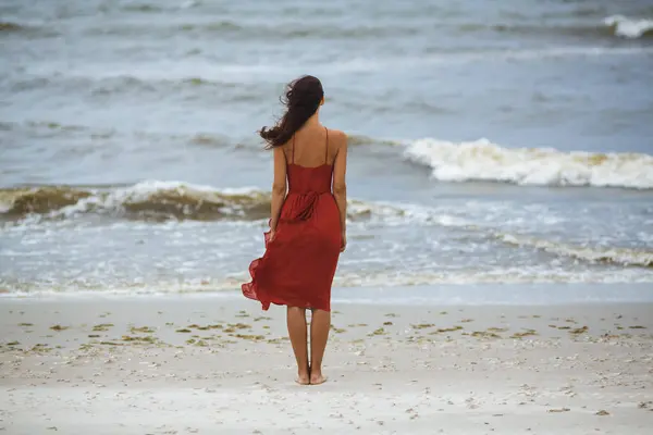 寒い風の強い天候に立っている赤いドレスの思慮深いビーチの女性 ストーミーウェーブ ストックフォト