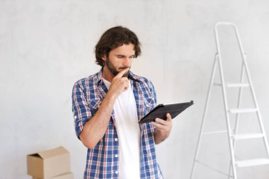 Tadilat nasıl yapılır. Boya rulosu tutan düşünceli genç bir kadının portresi, gri boyanmamış arka planda, merdivenli. Makale veya web sitesi için yüksek kaliteli fotoğraf