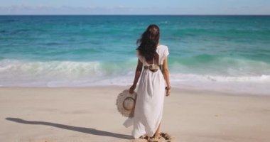 Beyaz elbiseli güzel, hassas sahil kadınının rüzgarlı havayla denize baktığı arka manzara, bohem bir kız. Yüksek kaliteli seyahat. Ağır çekimde ilham verici 4K görüntüler.