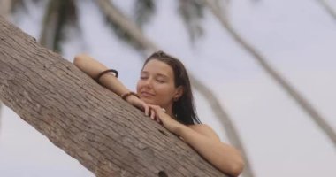 Siyah bikinili, bronzlaşmış bir kadın yaz plajında palmiye ağacının yanında dinleniyor.