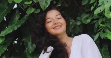 Mükemmel ciltli esmer kızın doğal saç bakımı portresi, doğal makyaj, egzotik bitkiler arasında ormanın önünde sağlıklı, parlak saçlarına dokunuyor. Yüksek kalite saç doğal kozmetik yavaş