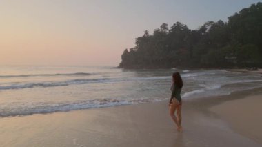 This amazing slow-motion 4K drone footage captures the breathtaking beauty of a slim girl in a bikini walking along the beach at sunrise. 