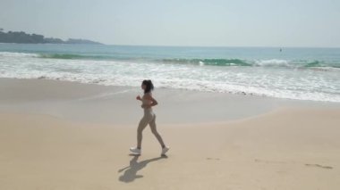 Aerial tracking shot young woman jogging on beautiful beach on summer morning. Active Asian female exercising outdoors running seashore. Concept of healthy lifestyle and sports. High quality 4k