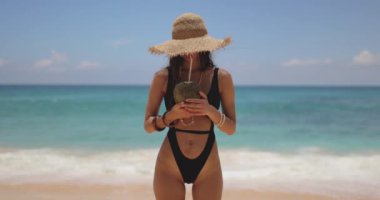 Relaxation in paradise during vacation of bikini woman in black swimsuit and straw hat on tropical beach near the ocean. Attractive brunette girl drinking coconut on a perfect sandy coast with blue
