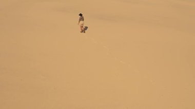 Çöl manzarasında kaybolan yorgun gezgin kadının yürüyüşü ve kurtarma arayışı. Kaşif kız sıcak bir günde kum tepeciğinde ayak izleri bırakıyor. Sinematik 4k drone görüntüleri
