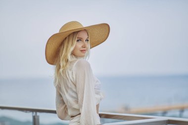 Yazlık plaj kıyafetiyle, deniz altyapısına karşı dışarıda dinlenen çarpıcı sarışın kadın. Moda modası geçmiş hasır şapka, beyaz bluz ve etek giyen, şık, romantik, genç bir bayan