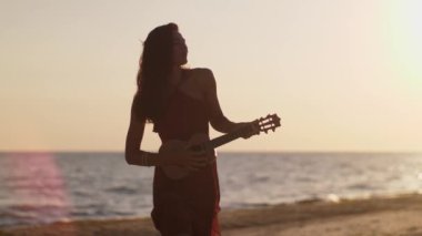 Yaz plajı titreşimleri. Gün batımında sahilde ukulele çalan siluetli bir kadın huzurlu ve sakin bir deniz havası yaratıyor. Sinema yavaş çekim 4k görüntü, reklam için tasarım ve sosyal