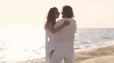 Beyaz elbiseli bir çift, güneşli bir plajda romantik bir öpücük paylaşıyor. Deniz kenarında güzel ve sevgi dolu bir anın tadını çıkarıyorlar..